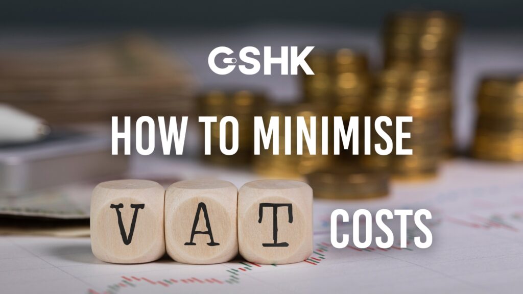 Cover image for 'How to Minimise VAT Costs,' featuring three wooden dice spelling 'VAT' with stacks of gold coins and financial charts blurred in the background.