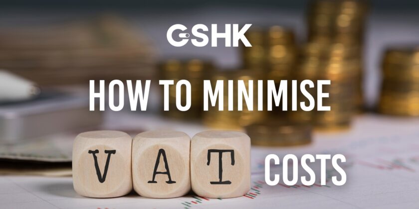 Cover Image For 'How To Minimise VAT Costs,' Featuring Three Wooden Dice Spelling 'VAT' With Stacks Of Gold Coins And Financial Charts Blurred In The Background.