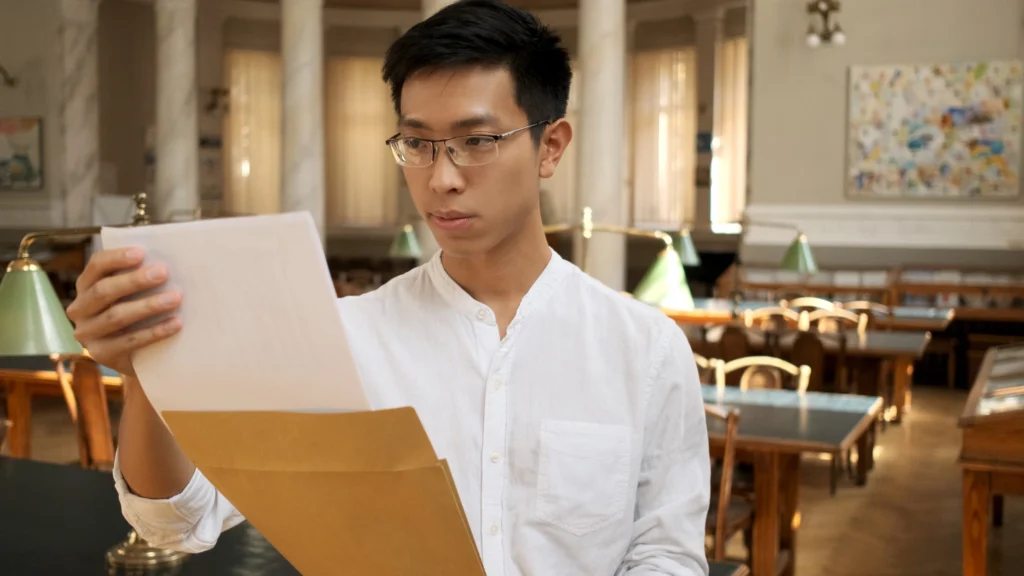 asian male student intently opening envelope with exam results university library