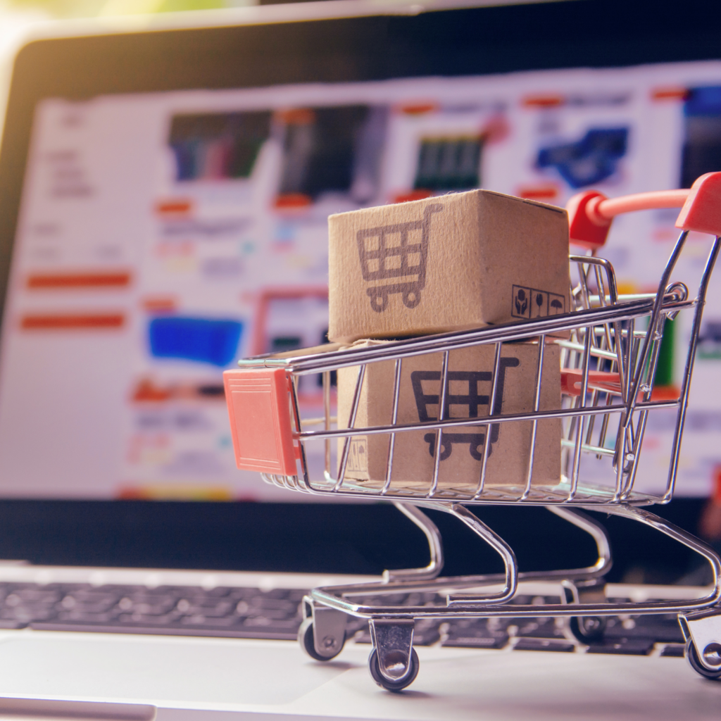 A miniature shopping cart on a laptop keyboard. Inside the cart are two small cardboard boxes with a shopping cart icon printed on them. The laptop screen in the blurred background shows a website with product listings.
