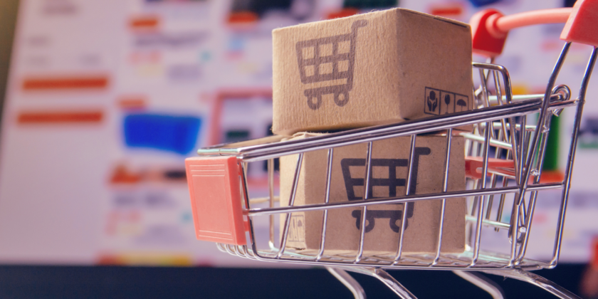 A Miniature Shopping Cart On A Laptop Keyboard. Inside The Cart Are Two Small Cardboard Boxes With A Shopping Cart Icon Printed On Them. The Laptop Screen In The Blurred Background Shows A Website With Product Listings.