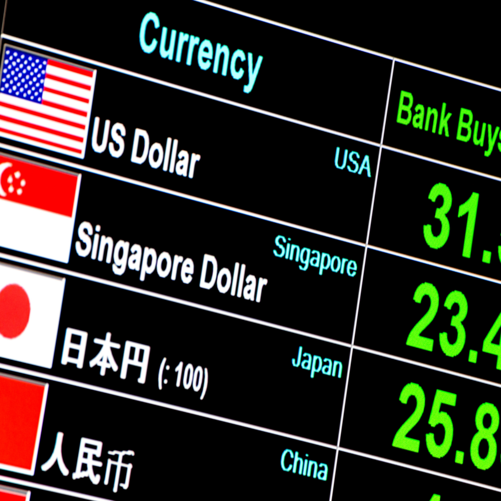 A close-up photograph of a digital currency exchange rate board with a black background. The screen displays a list of currencies with their national flags and abbreviations, including the US Dollar (USA), Singapore Dollar (Singapore), Japanese Yen (Japan), and Chinese Yuan (China). This image indicates the risk of FX exposure.
